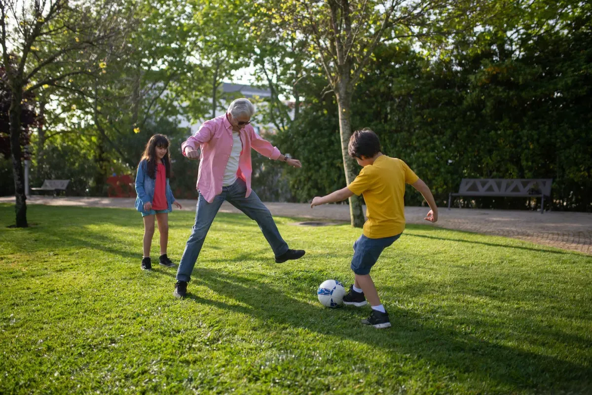 Un abuelo juega fútbol en un parque con su nieta y su nieto.